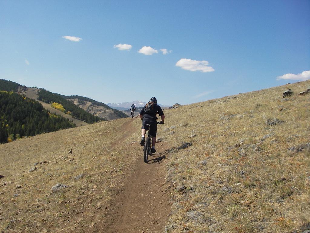 Two mountain bikers ride along a dirt trail on a hillside, surrounded by open grassland and distant mountains under a blue sky with a few clouds. Clear Creek mountain bike trail.