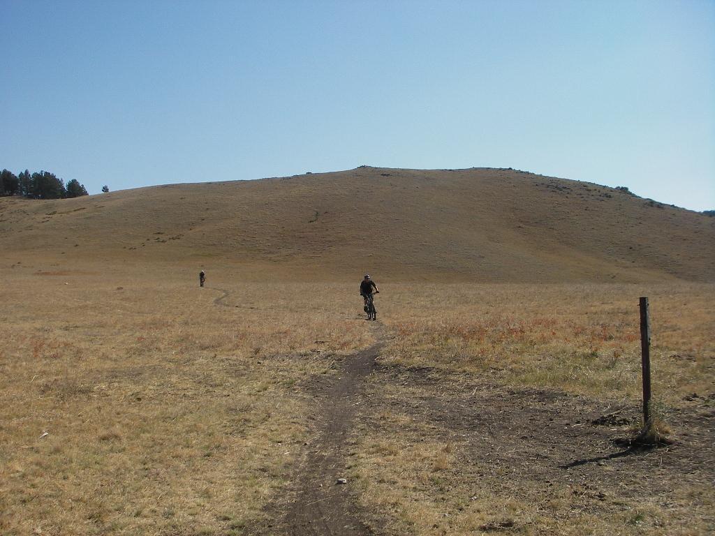 Two cyclists riding on a dirt path through a vast, dry grassland, with rolling hills in the background under a clear blue sky. Clear Creek mountain bike trail.