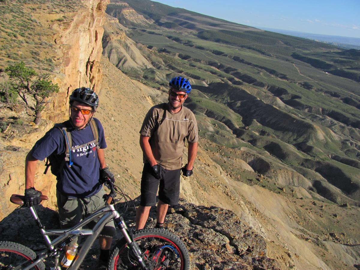 Two mountain bikers standing near the edge of a rocky cliff, overlooking a vast, rugged landscape with rolling hills and valleys in the background. One biker is holding a mountain bike and smiling, while the other is standing beside him, both wearing helmets and casual outdoor clothing. The scene captures a sense of adventure and the beauty of outdoor recreational activities. Palisade Rim mountain bike trail.