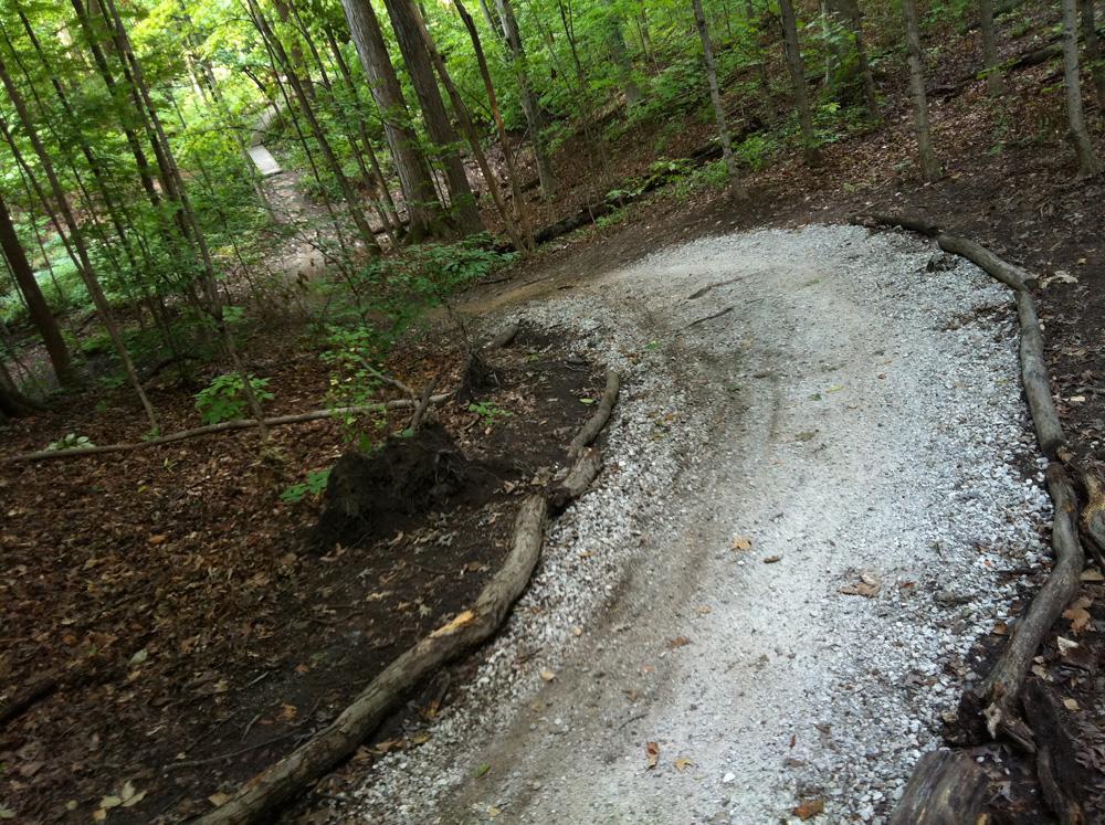 A winding gravel trail through a wooded area, surrounded by lush green trees and scattered leaves. The path curves gently, flanked by logs and underbrush, inviting walkers or cyclists to explore the natural landscape. Franke Park mountain bike trail.