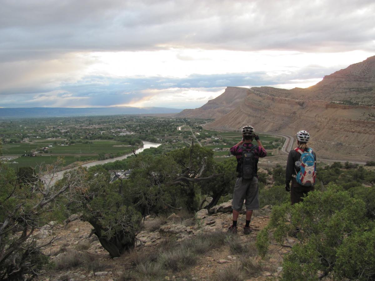 Two individuals wearing helmets stand on a rocky overlook, gazing at a scenic vista of rolling hills and a winding river below. The landscape features green fields and small towns, under a cloudy sky with hints of sunlight peeking through. Palisade Rim mountain bike trail.