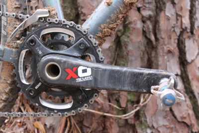 Close-up of a bicycle's crankset and pedals, showing a muddy chainring and a crank arm with a logo, positioned against a textured tree trunk background.