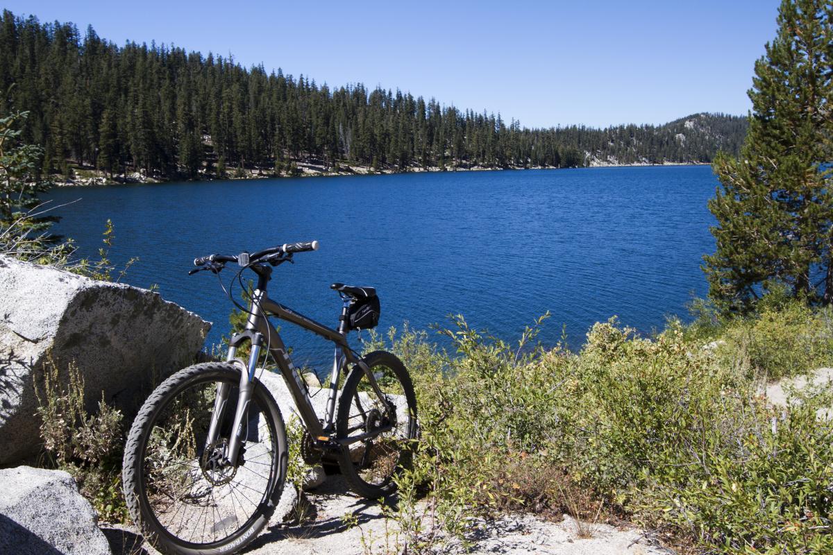 A mountain bike resting on a rock near a tranquil lake, surrounded by lush greenery and coniferous trees under a clear blue sky. Flume Trail mountain bike trail.