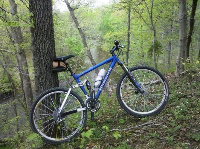 A blue mountain bike parked on a hillside surrounded by lush green trees and vegetation, near a gentle stream. A water bottle is attached to the bike, indicating readiness for an outdoor adventure. Wincopin (Savage) Trail mountain bike trail.