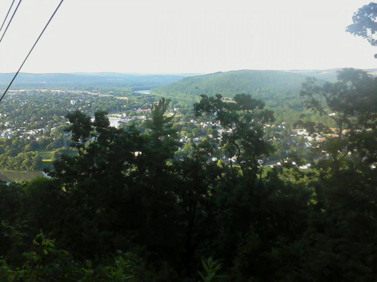 A scenic view from a hillside overlooking a lush green valley and a small town, with trees in the foreground and distant mountains on the horizon under a clear sky. Power lines are visible at the top of the image. Round Top mountain bike trail.