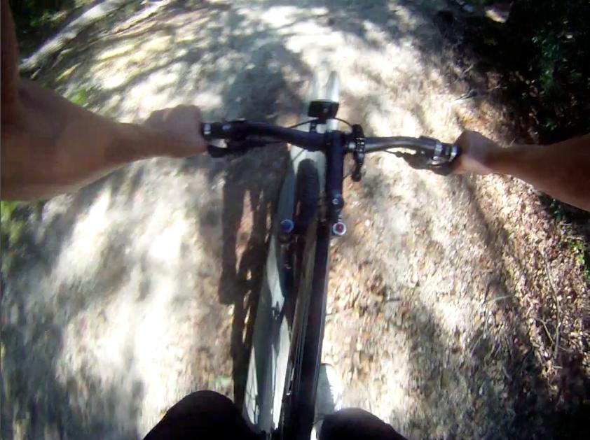 A top-down view of a mountain bike in motion on a dirt trail, with hands gripping the handlebars and sunlight filtering through surrounding trees. Allaire State Park mountain bike trail.