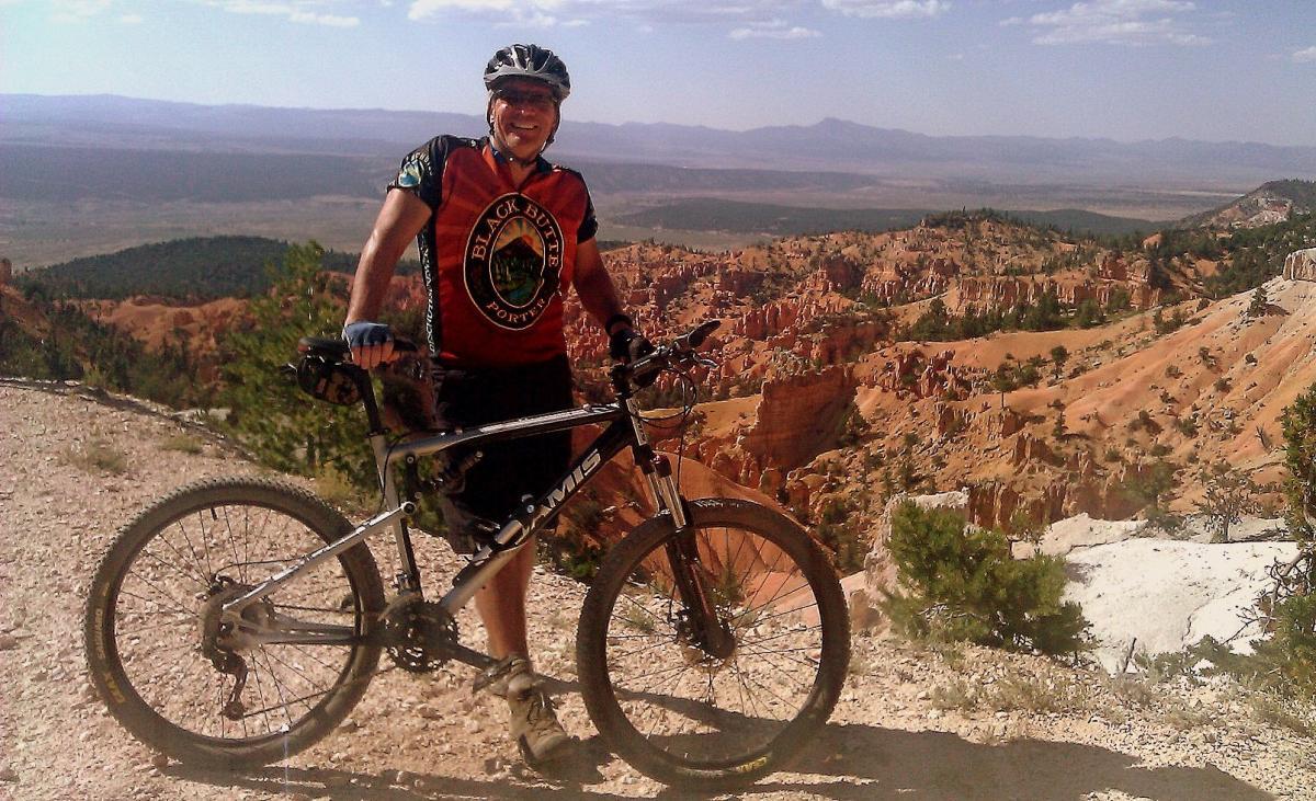 A person in a cycling outfit stands next to a mountain bike on a trail with stunning views of a rugged landscape. The background features layered rock formations and greenery, with mountains visible in the distance under a clear blue sky. The individual is smiling, showcasing a sense of adventure and enjoyment in an outdoor setting. Thunder Mountain mountain bike trail.