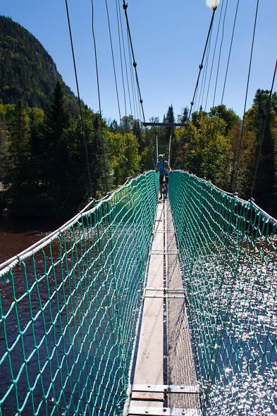 A person walking on a suspension bridge with a green netting safety barrier, surrounded by lush trees and mountains under a clear blue sky. The bridge spans over a shimmering river below. Vallee Bras Du Nord Secteur Shannahan mountain bike trail.
