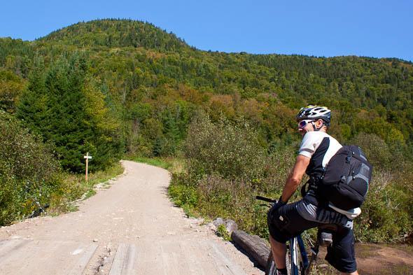 A cyclist in athletic gear sits on a mountain bike at a crossroads on a dirt path, surrounded by lush green trees and a hillside in the background. The scene is set on a clear day with a blue sky, indicating a perfect day for outdoor adventure. A signpost is visible on the left, guiding the way through the natural landscape. Vallee Bras Du Nord Secteur Shannahan mountain bike trail.