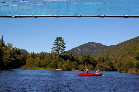A bright blue sky above a serene river with two people paddling in a red canoe. In the background, a suspension bridge is visible, along with lush green trees and mountains. The scene captures a peaceful moment in nature, showcasing the beauty of the outdoors. Vallee Bras Du Nord Secteur Shannahan mountain bike trail.