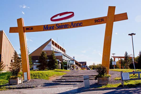 Entrance sign to Mont Sainte-Anne, a ski resort, featuring a large wooden arch with the name "Mont Sainte-Anne" prominently displayed. In the background, the resort's main building can be seen, along with surrounding greenery and trees. Mont-Sainte-Anne mountain bike trail.