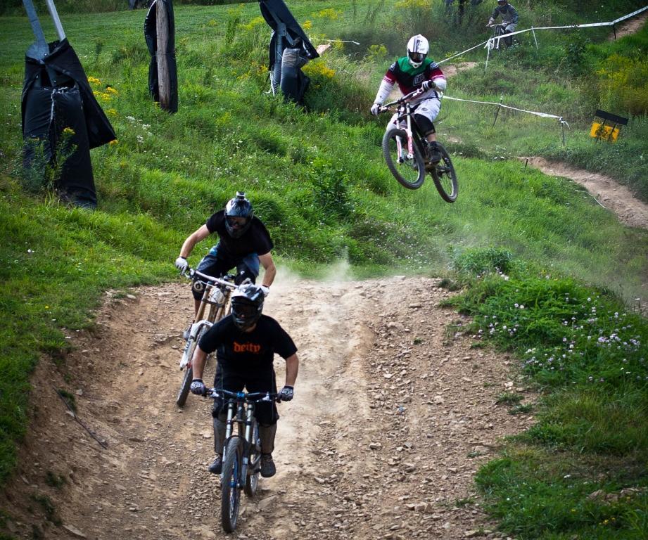 A group of three mountain bikers navigate a dirt track surrounded by green grass and wildflowers. One rider is airborne, performing a jump, while the others are positioned on the ground. Dust is kicked up from the tires, capturing the excitement of the scene in a natural setting. Seven Springs mountain bike trail.