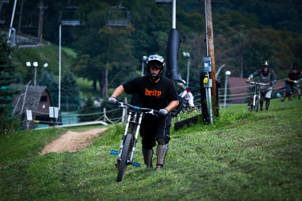 A group of mountain bikers is riding on a grassy slope at a bike park, with a ski lift in the background. One rider, wearing a black t-shirt and protective gear, is in the foreground, focusing on navigating the terrain. The landscape features trees and a few buildings, with more bikers visible in the background, enjoying the descent. Seven Springs mountain bike trail.