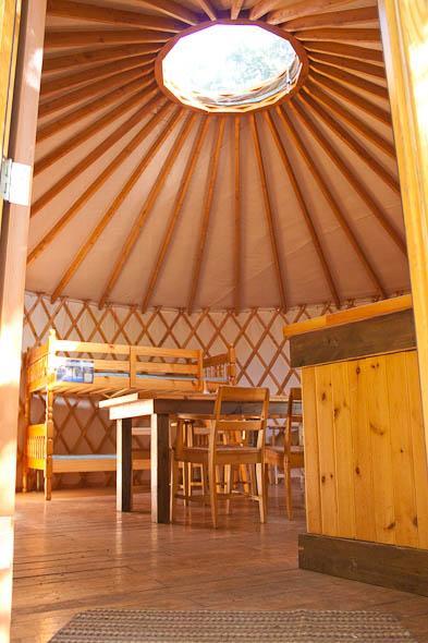 Interior of a yurt featuring wooden beams and a circular skylight. The space includes bunk beds, a wooden dining table, and several chairs, all on a wooden floor. The walls are adorned with traditional yurt latticework. Vallee Bras Du Nord Secteur Shannahan mountain bike trail.