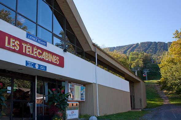 The image shows a modern building marked "Les Télécabines," likely a ski lift station. The structure features large windows and a sign above the entrance. In the background, a hillside is visible, with ski lifts and greenery indicating a mountainous area. The scene is bathed in natural light, reflecting a clear, sunny day. Mont Sainte-anne mountain bike trail.