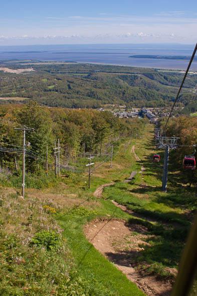 A scenic view from a ski lift overlooking a mountainside with greenery, leading down to a valley and a body of water in the distance. The sky is clear with a few clouds, and the ski lift towers and cables are visible along the slope. Mont Sainte-anne mountain bike trail.