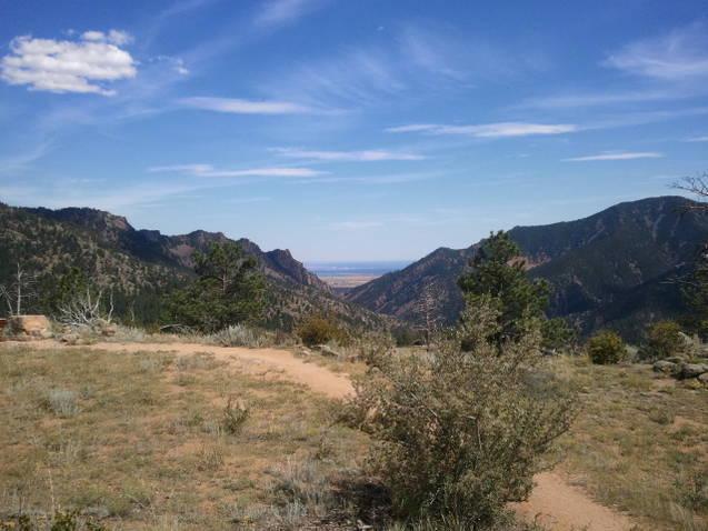 A scenic view of a mountainous landscape with a clear blue sky, featuring a winding dirt path in the foreground, surrounded by vegetation. The valley below is visible, flanked by rugged mountains on either side, creating a peaceful and natural environment. Walker Ranch mountain bike trail.