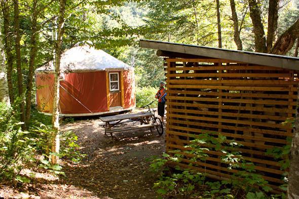 A peaceful campsite scene featuring a circular orange yurt nestled among trees, with a wooden shelter nearby and a picnic table in the foreground. Sunlight filters through the leaves, creating a tranquil atmosphere. A person is visible in the background, engaged in an activity. Vallee Bras Du Nord Secteur Shannahan mountain bike trail.