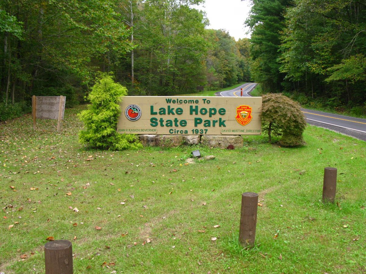 Welcome sign for Lake Hope State Park, featuring green lettering on a wooden structure, surrounded by trees and grassy areas, with a road visible in the background. The sign notes the park's establishment date as 1937 and includes various insignia. Lake Hope State Park mountain bike trail.
