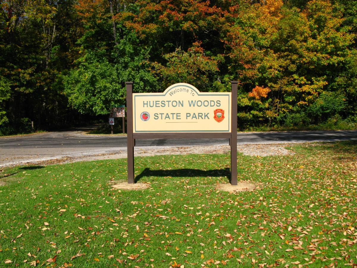 A welcome sign for Hueston Woods State Park, featuring the park's name prominently displayed. The sign is set against a backdrop of trees displaying autumn foliage, with a grassy area in the foreground and a paved road visible. Hueston Woods State Park mountain bike trail.