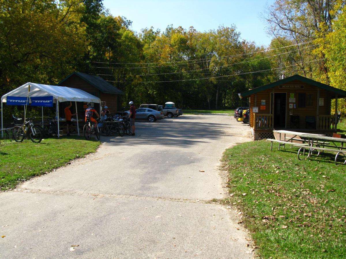 A view of a bike rental area with a tent and a wooden building. Several parked bicycles are in front of the tent, and people are interacting around the area. The scene is set in a natural environment with trees displaying autumn foliage. A small parking lot is visible with cars parked nearby, and a picnic table is situated on the grass. The weather appears clear and sunny. Hueston Woods State Park mountain bike trail.