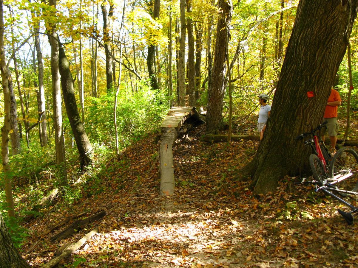 A forested trail with autumn foliage, featuring a wooden bridge positioned slightly uphill. Two people are visible in casual clothing, one wearing a helmet, while a bicycle is leaning against a tree nearby. The ground is covered with fallen leaves, and the scene is bright with natural light filtering through the trees. Hueston Woods State Park mountain bike trail.