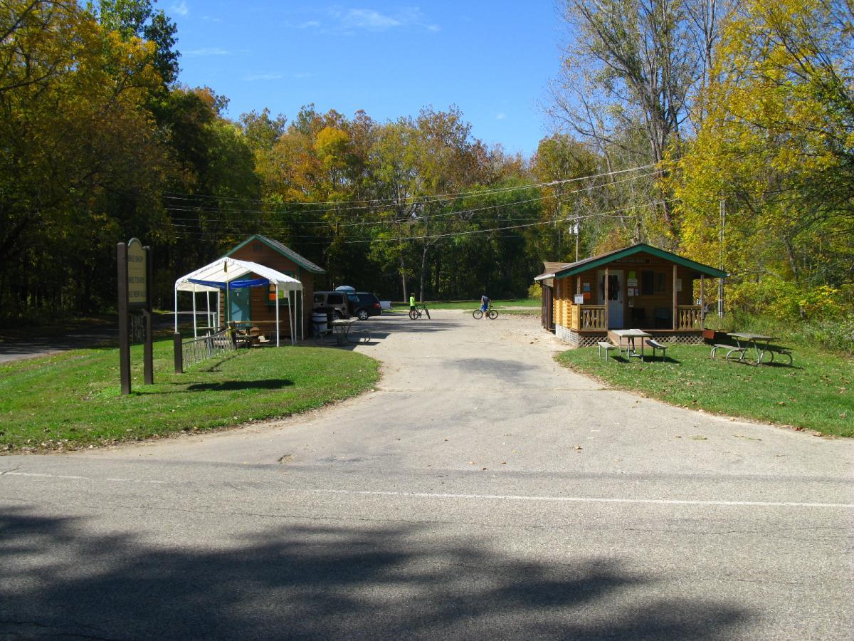 Image of a park entrance with two small wooden buildings on either side of a curved road. One building has a porch and a sign nearby, while the other is covered by a white canopy. There are picnic tables in the grass, and a person on a bicycle can be seen in the background, surrounded by trees displaying autumn foliage under a clear blue sky. Hueston Woods State Park mountain bike trail.