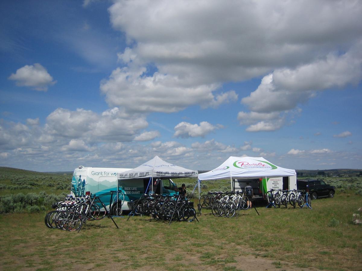 A mobile bike shop setup in an open field featuring a van and two tents, surrounded by several bicycles on display. The sky is filled with fluffy clouds, and the area is mostly green with sparse vegetation.