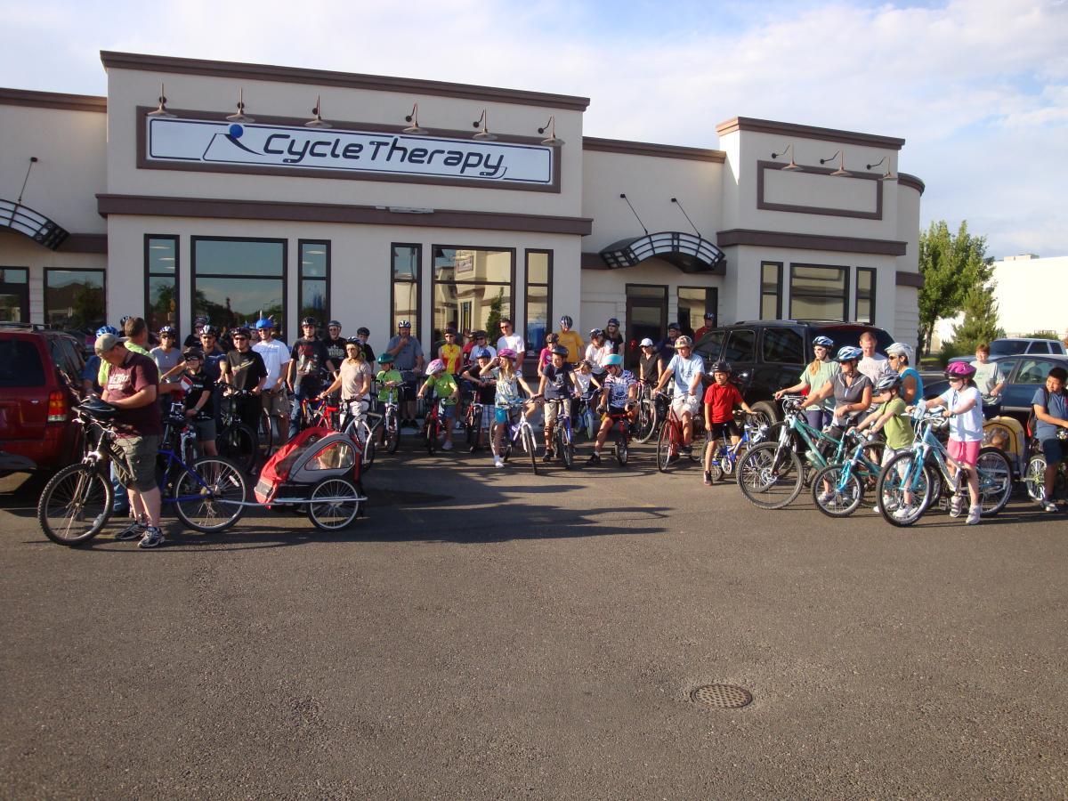 A group of people, including children and adults, gathered outside a bike shop named "Cycle Therapy." Many participants are wearing helmets and are standing alongside their bicycles, with some people using a bike trailer. The scene portrays a community event or bike ride initiative, set against a clear blue sky.