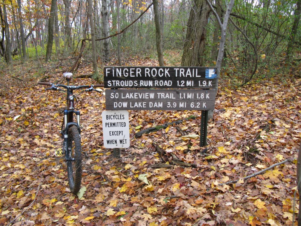 A mountain bike parked next to a trail sign indicating "Finger Rock Trail" with distances to nearby routes. The forest floor is covered in colorful autumn leaves, and the background features trees in fall foliage. The sign includes trail names and distance measurements, with a note about bicycle use restrictions when wet. Strouds Run State Park mountain bike trail.