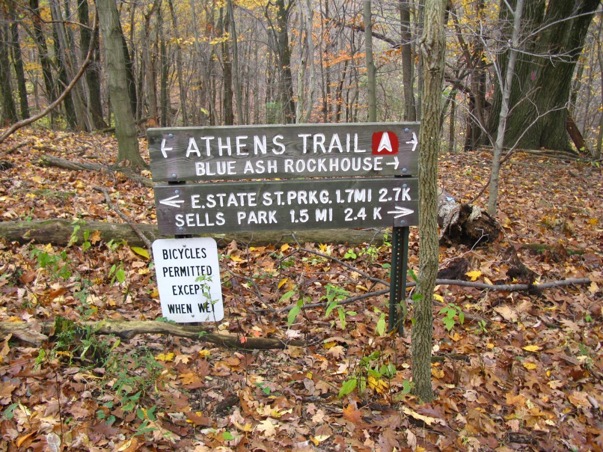 A wooden trail sign indicating directions to Athens Trail and Blue Ash Rockhouse, along with distances to nearby locations such as E. State St. Park and Sells Park. The ground is covered with fallen leaves, and a separate sign states that bicycles are permitted except when wet. Strouds Run State Park mountain bike trail.