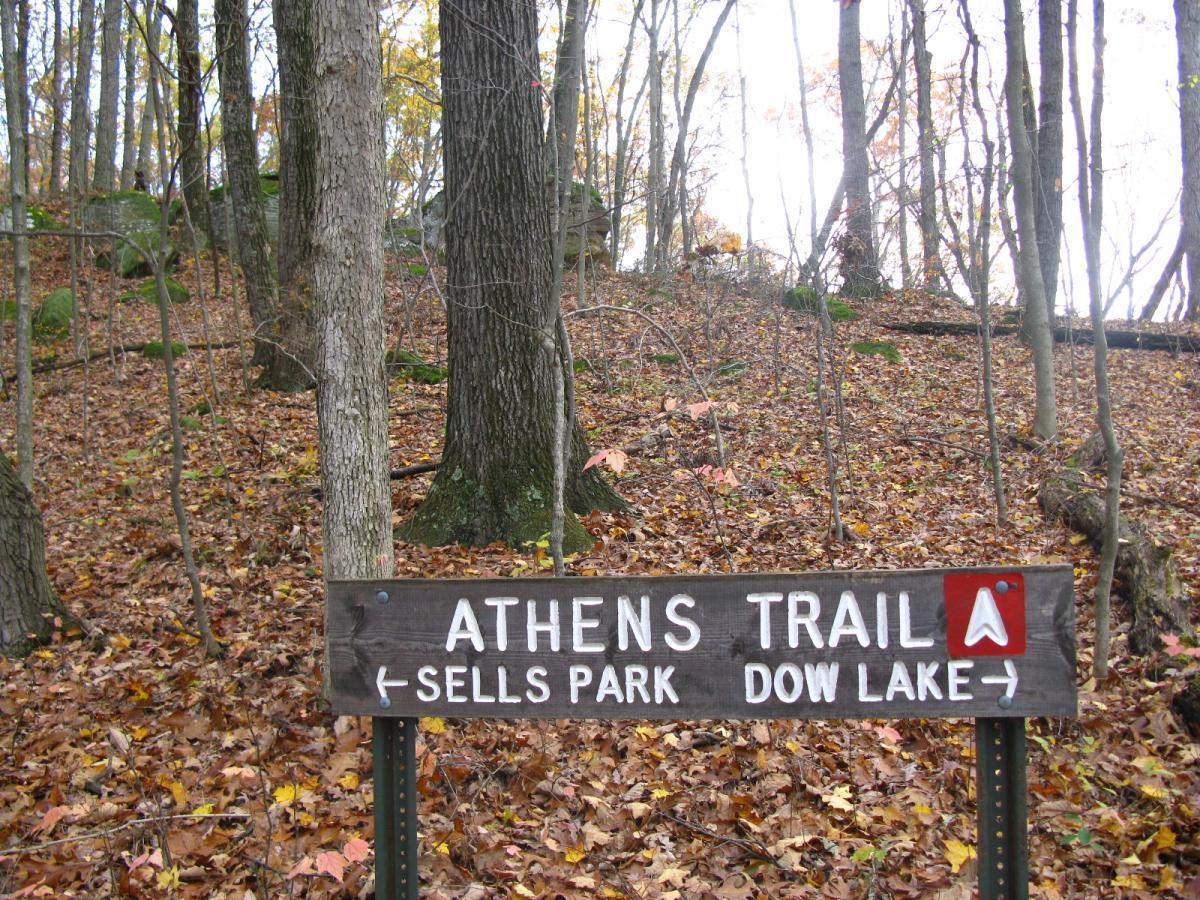 Wooden trail sign indicating the Athens Trail with directional arrows pointing towards Sells Park and Dow Lake, surrounded by a forested area with autumn leaves scattered on the ground. Strouds Run State Park mountain bike trail.