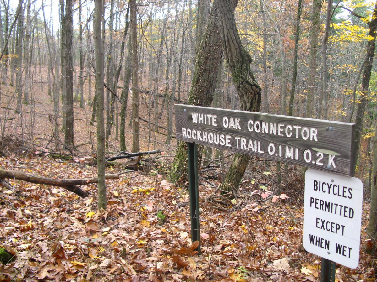 A wooded area in autumn with fallen leaves covering the ground. A wooden sign indicates the "White Oak Connector" and "Rockhouse Trail" with distance markers of 0.1 miles and 0.2 kilometers. Another sign below states that bicycles are permitted except when the trail is wet. Strouds Run State Park mountain bike trail.