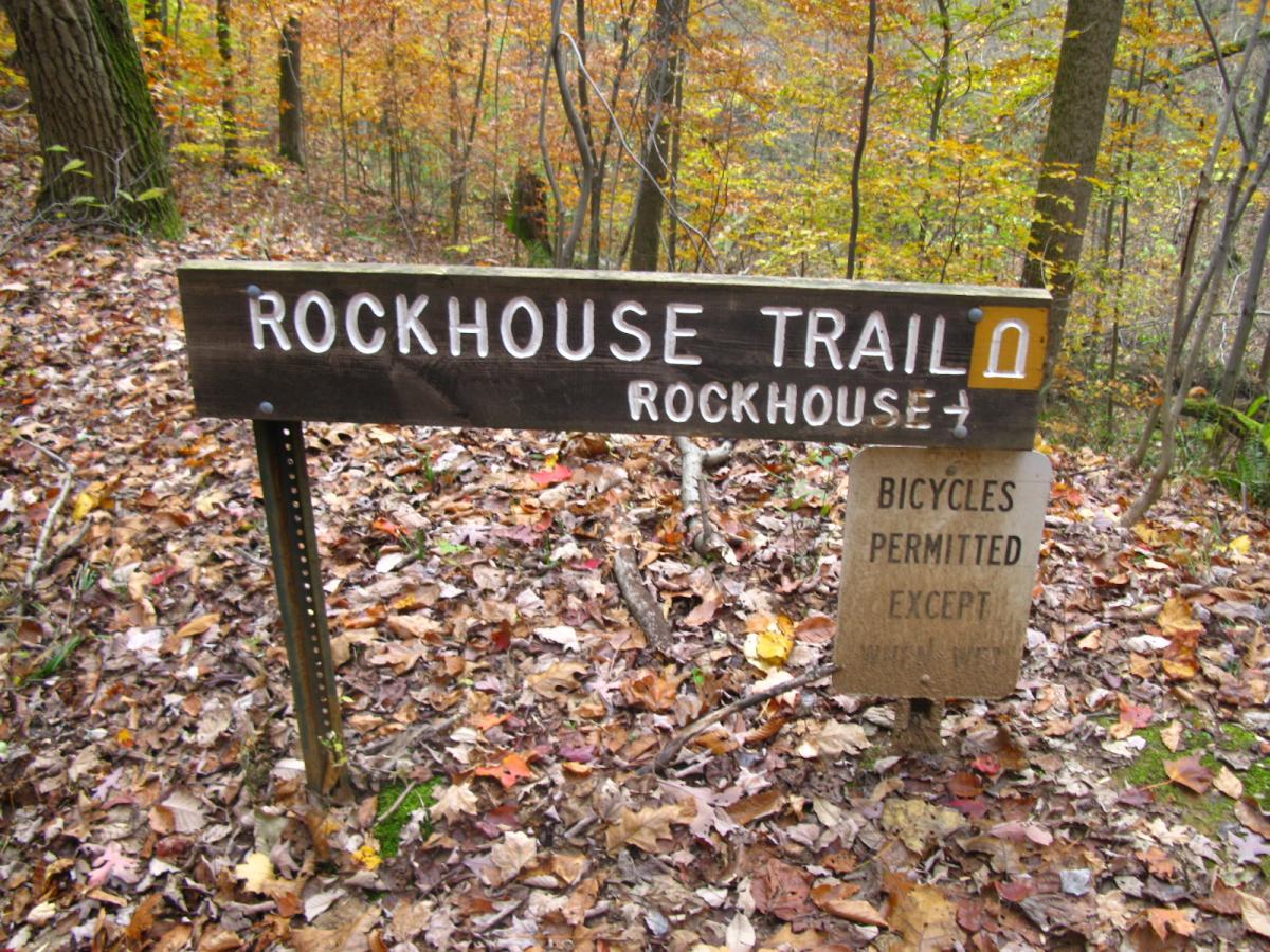 Wooden trail sign for "Rockhouse Trail" with text indicating bicycles are permitted, set in a forested area with autumn leaves covering the ground. Strouds Run State Park mountain bike trail.