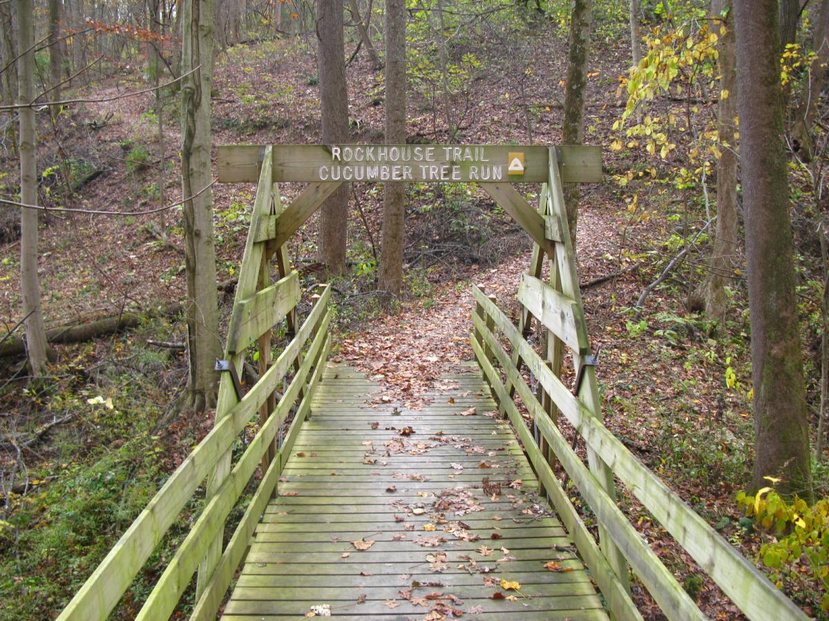 Wooden footbridge leading to Rockhouse Trail, marked with a sign reading "Rockhouse Trail Cucumber Tree Run," surrounded by trees and fallen leaves on the forest floor. Strouds Run State Park mountain bike trail.
