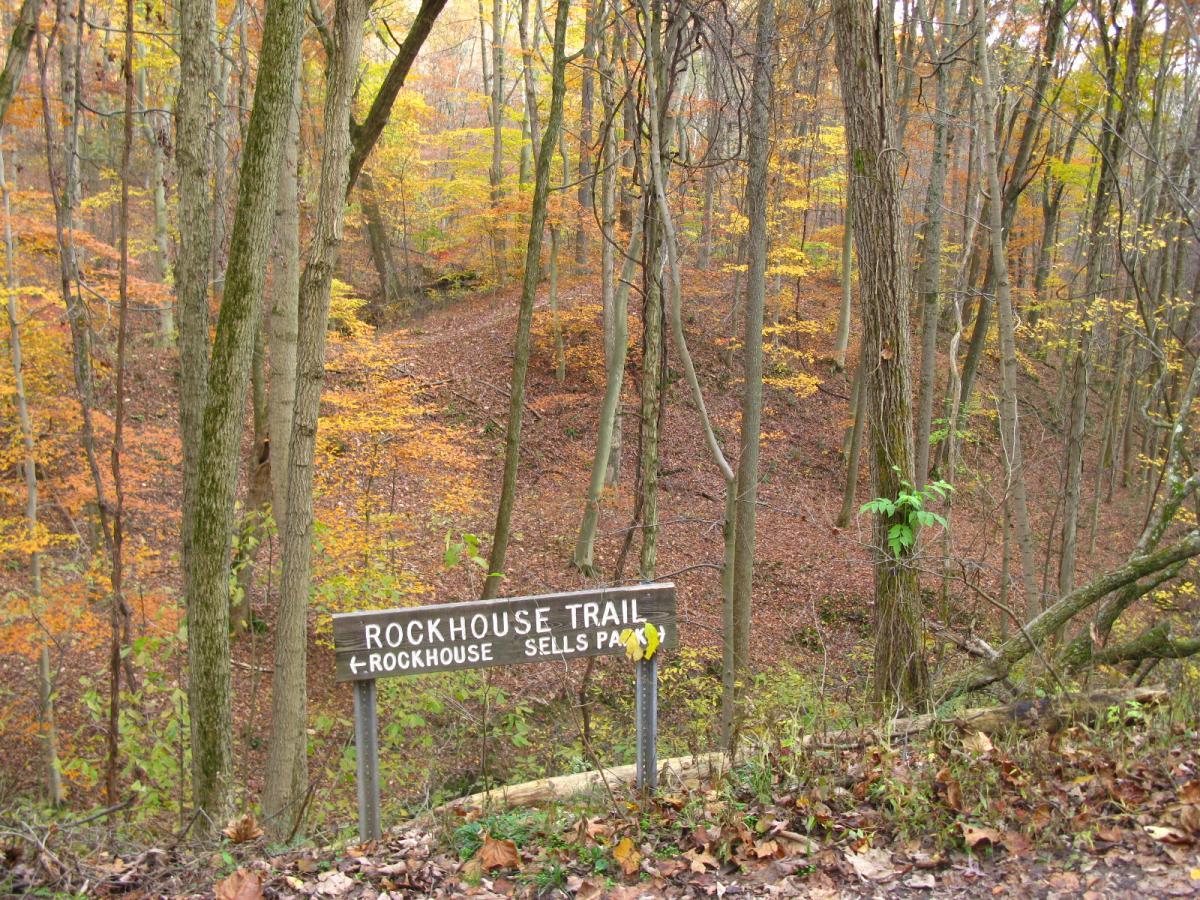 A wooden sign labeled "Rockhouse Trail" pointing left towards Rockhouse and Sells Park, surrounded by a colorful autumn forest with orange and yellow leaves, and a winding trail in the background. Strouds Run State Park mountain bike trail.