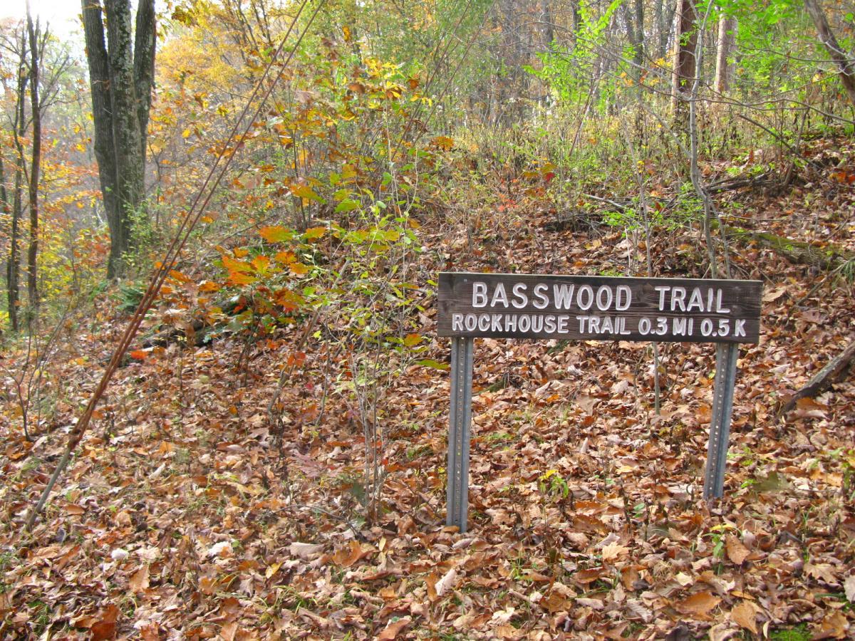 Wooden trail sign indicating "Basswood Trail" and "Rockhouse Trail," with information about trail distances, surrounded by autumn foliage and fallen leaves. Strouds Run State Park mountain bike trail.