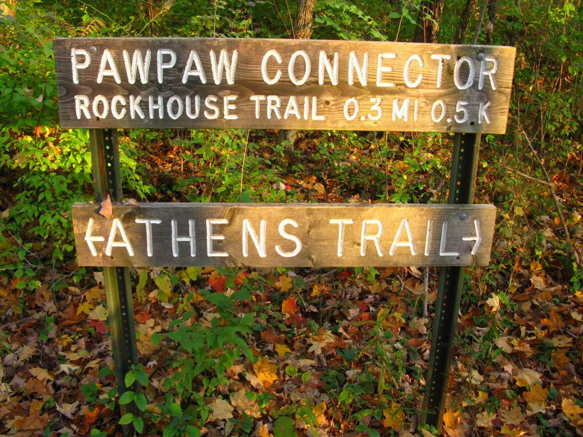 Wooden trail sign indicating the Pawpaw Connector and Rockhouse Trail with distances of 0.3 miles and 0.5 kilometers, along with directions to Athens Trail, surrounded by autumn foliage and greenery. Strouds Run State Park mountain bike trail.