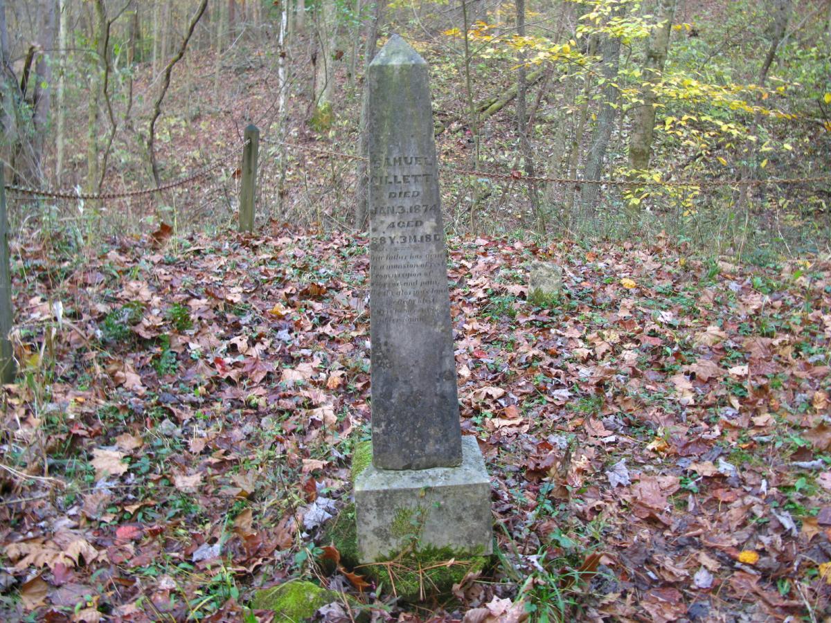 A weathered gravestone stands in a wooded area, surrounded by fallen leaves and greenery. The stone is engraved with the name Samuel Tilley, along with dates indicating his birth and death in the 19th century. The setting features a natural landscape with trees and a chain-link fence in the background, suggesting a serene yet somber atmosphere. Strouds Run State Park mountain bike trail.