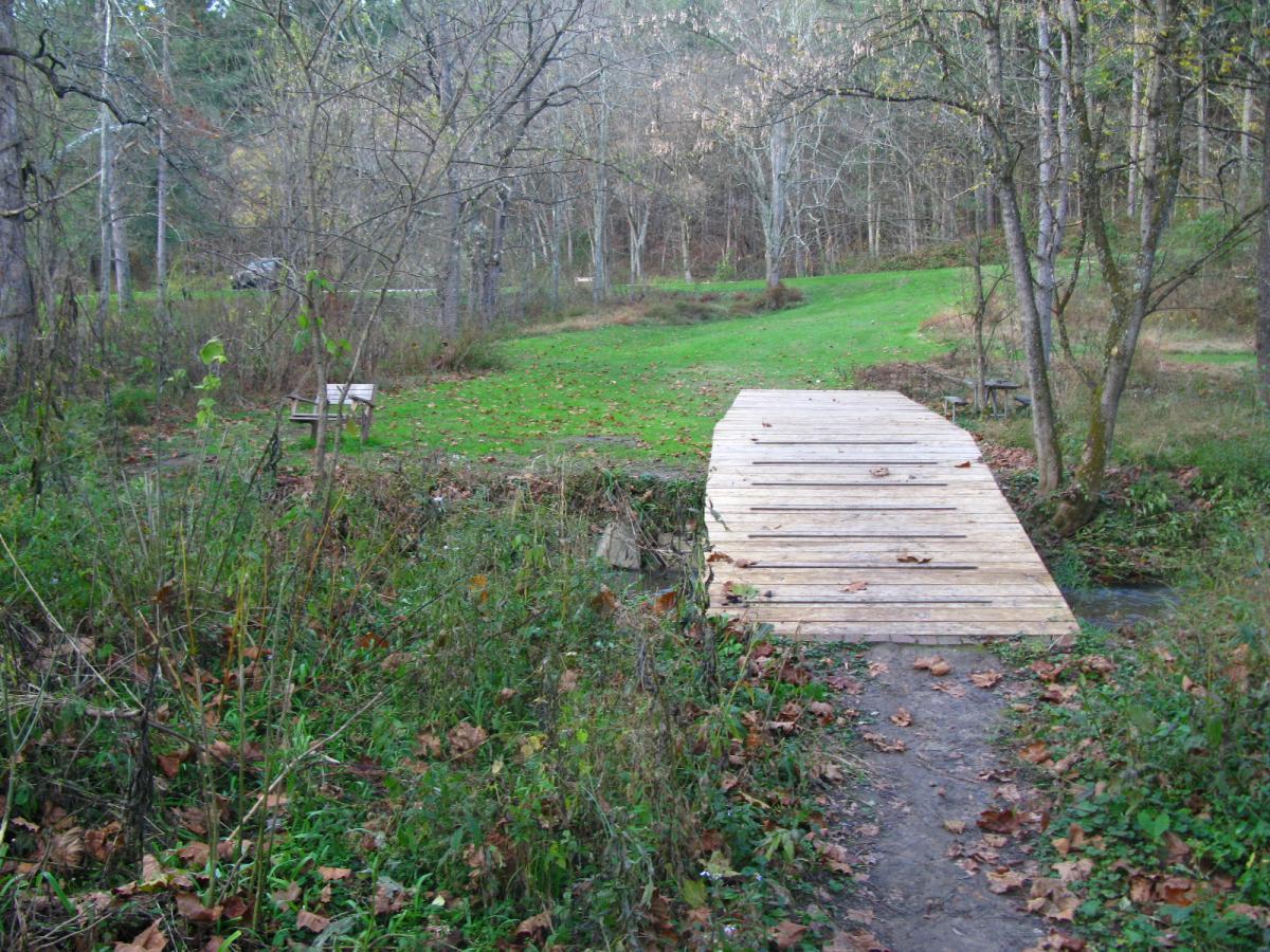 A wooden bridge crossing a small stream, surrounded by a natural, wooded landscape with trees and fallen leaves. A grassy area is visible in the background, along with a wooden bench partially obscured by foliage. Strouds Run State Park mountain bike trail.