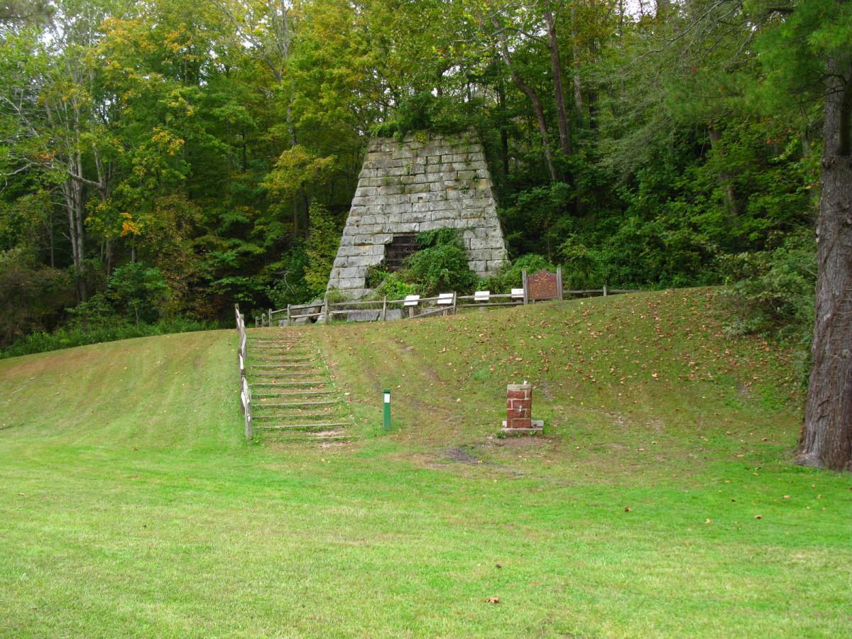 A stone monument sits at the edge of a grassy hill, surrounded by trees. A set of wooden stairs leads from the grassy area to the base of the monument. Benches are positioned nearby for visitors, and a small sign can be seen to the right of the monument, indicating points of interest and history. Lake Hope State Park mountain bike trail.
