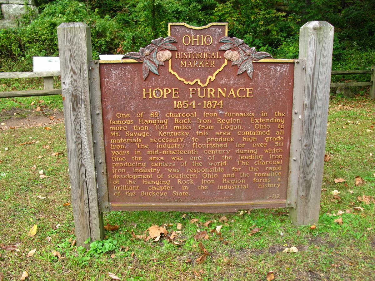 A bronze historical marker titled "Hope Furnace 1854-1874," located in Ohio. The plaque provides information about the Hope Furnace, one of the 69 charcoal iron furnaces in the Hanging Rock Iron Region. The text discusses its significance in producing high-grade iron and its role in the industrial development of southern Ohio during the mid-nineteenth century. The marker is set against a backdrop of trees and natural vegetation. Lake Hope State Park mountain bike trail.