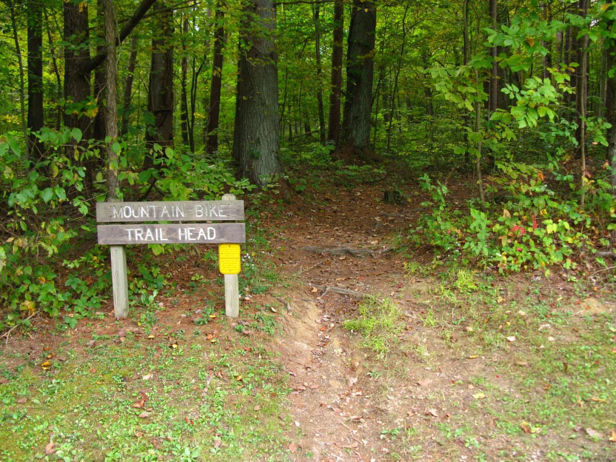 Sign indicating the entrance to a mountain bike trail head, surrounded by lush green trees and foliage, with a dirt path leading into the woods. Lake Hope State Park mountain bike trail.