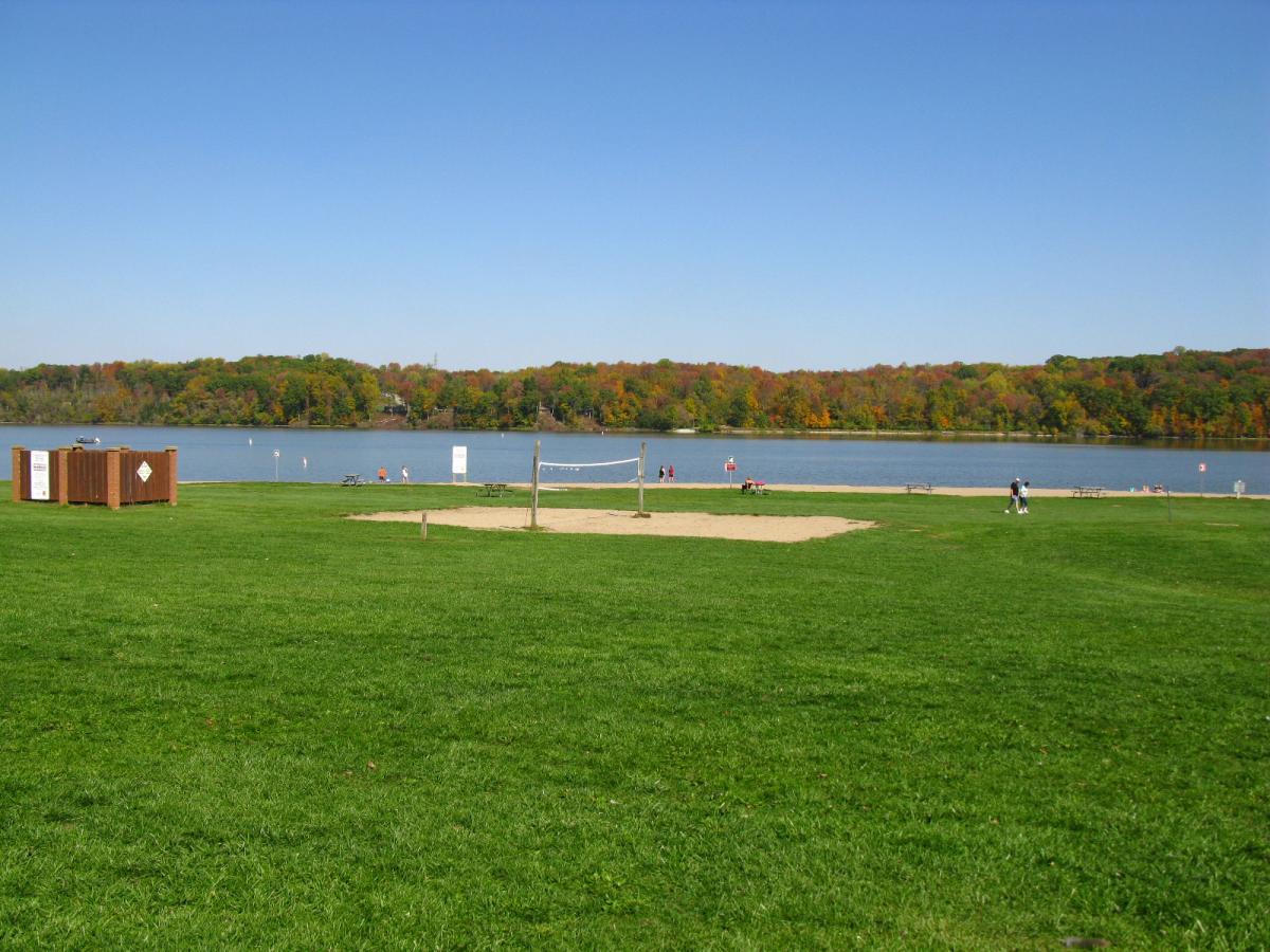 A sunny lakeside park featuring a sandy volleyball court, green grass, and colorful trees in the background during autumn. There are a few people near the water, and a wooden structure nearby. The clear blue sky enhances the serene atmosphere. Hueston Woods State Park mountain bike trail.