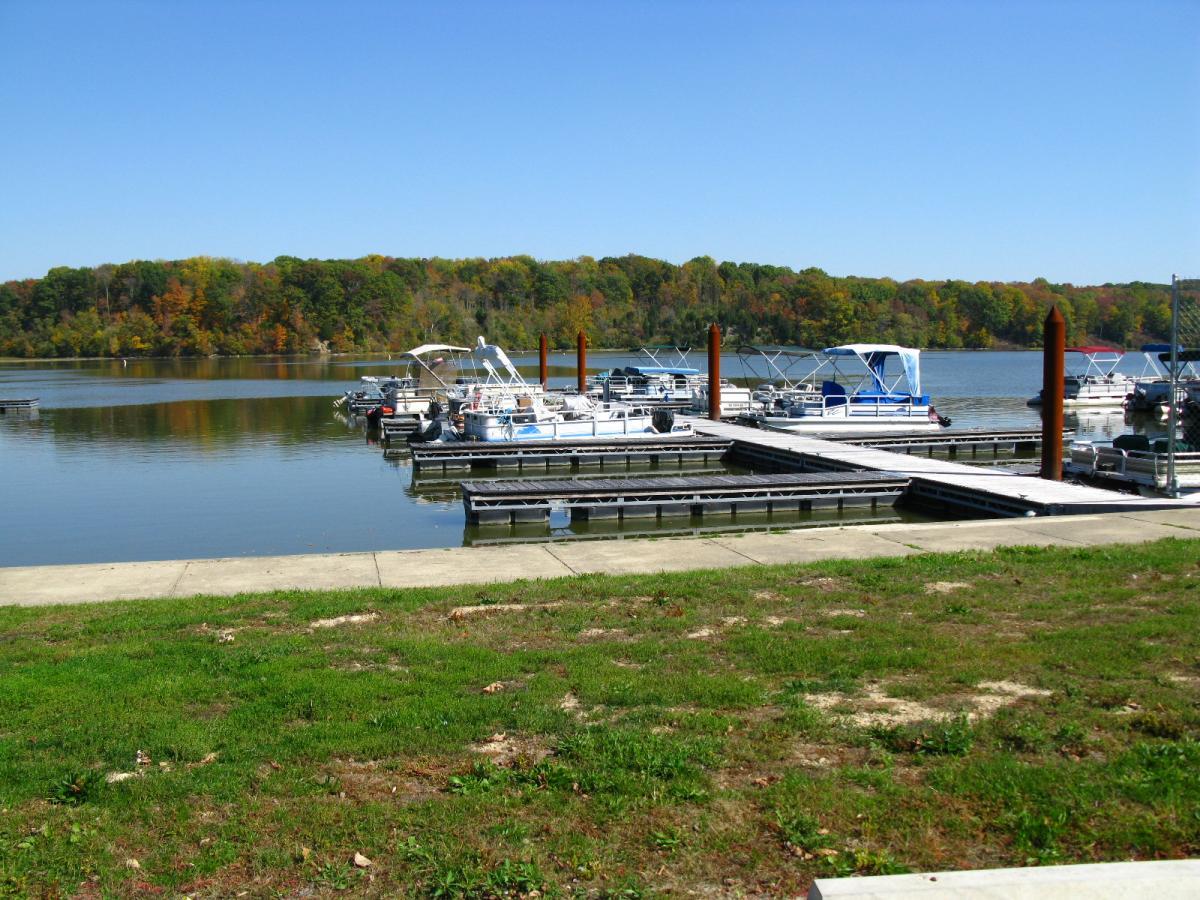 A serene lakeside scene featuring a dock with several boats moored. The water is calm, reflecting the clear blue sky above. In the background, a forested area shows vibrant autumn foliage with shades of orange and green. The grassy foreground includes a paved area leading to the dock, suggesting a peaceful recreational spot. Hueston Woods State Park mountain bike trail.