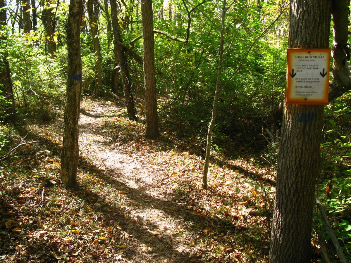 A winding dirt path through a wooded area, surrounded by green trees and scattered autumn leaves. A sign mounted on a tree provides trail information and safety reminders. Sunlight filters through the foliage, creating a serene and inviting atmosphere. Hueston Woods State Park mountain bike trail.