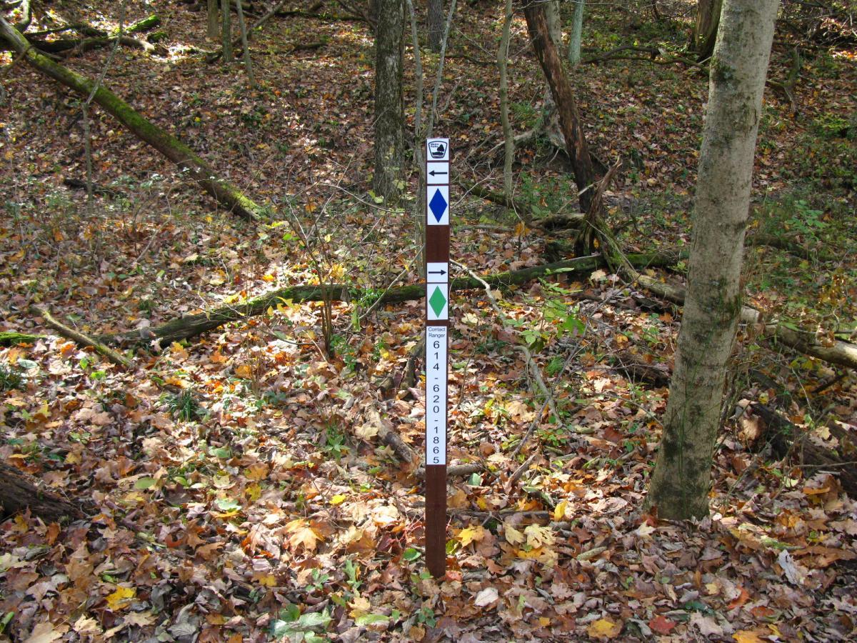 A trail marker post in a wooded area, surrounded by fallen leaves. The post displays a series of colored diamonds—blue, green, and white—along with a contact number for the local ranger. Chestnut Ridge mountain bike trail.