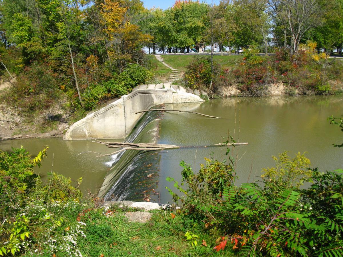 A serene landscape featuring a small concrete dam spanning a gently flowing river. The water cascades over the dam, creating a tranquil waterfall effect. Surrounding the river is lush greenery and trees displaying autumn foliage, with hints of yellow, orange, and red. A walking path and additional trees are visible in the background, enhancing the peaceful outdoor scene. Van  Buren mountain bike trail.