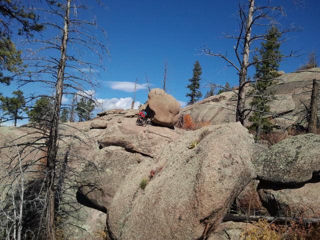 A landscape featuring large, rugged boulders and scattered pine trees under a bright blue sky with a few clouds. One of the boulders is prominently positioned, and the terrain appears rocky and uneven, suggesting an outdoor hiking or climbing environment. Buffalo Creek mountain bike trail.