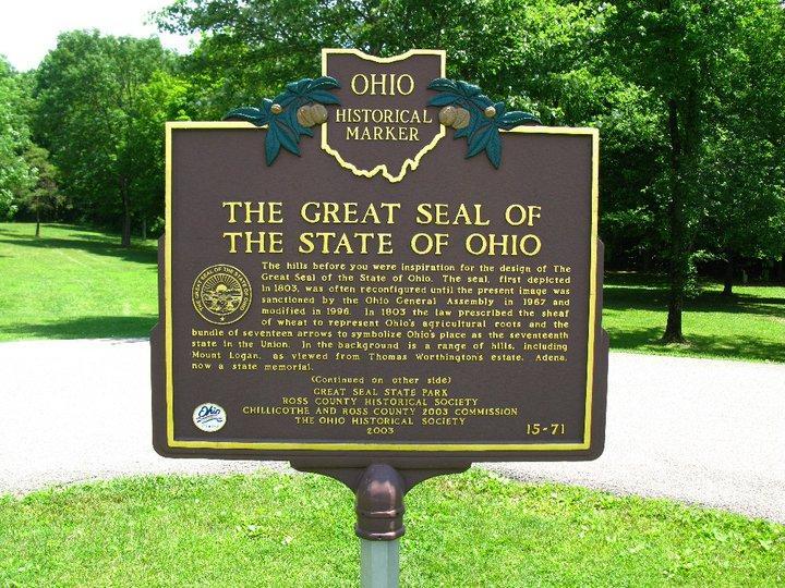 A historical marker in a park, featuring information about "The Great Seal of the State of Ohio." The sign is brown with yellow lettering, detailing the origins of the seal, its design inspiration from local hills, and its legislative history. Surrounding the marker are lush green trees and grass, indicating a natural setting. Great Seal State Park mountain bike trail.